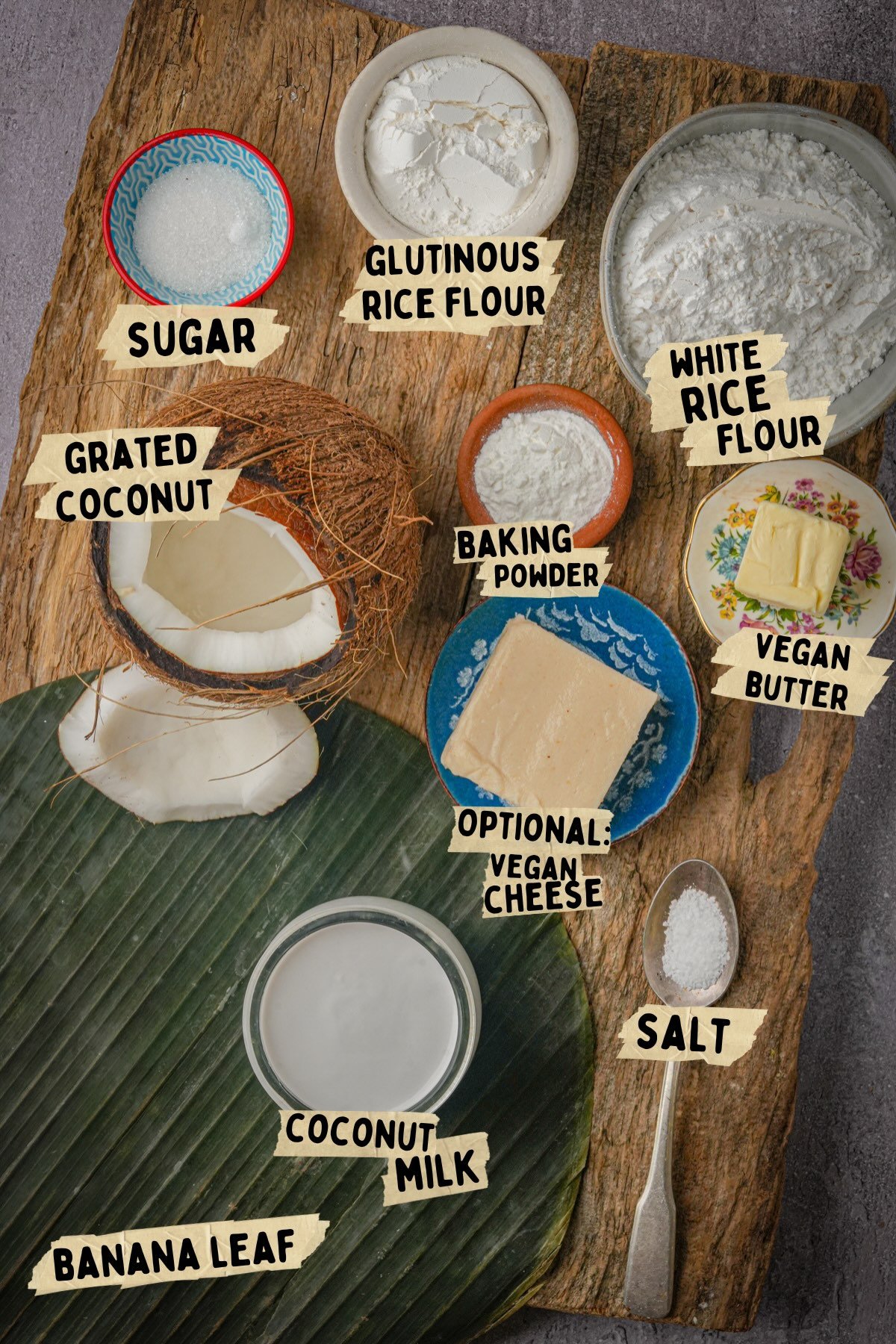 Ingredients for a vegan bibingka recipe displayed on a wooden surface, including labeled bowls of sugar, glutinous rice flour, white rice flour, baking powder, vegan butter, grated coconut, optional vegan cheese, salt, coconut milk, and a banana leaf.