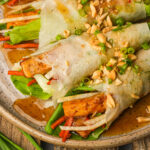 Fresh lumpiang sariwa rolls filled with lettuce, carrots, tofu, and fresh herbs, topped with chopped peanuts and green onions, served with a drizzle of brown sauce on a rustic plate.