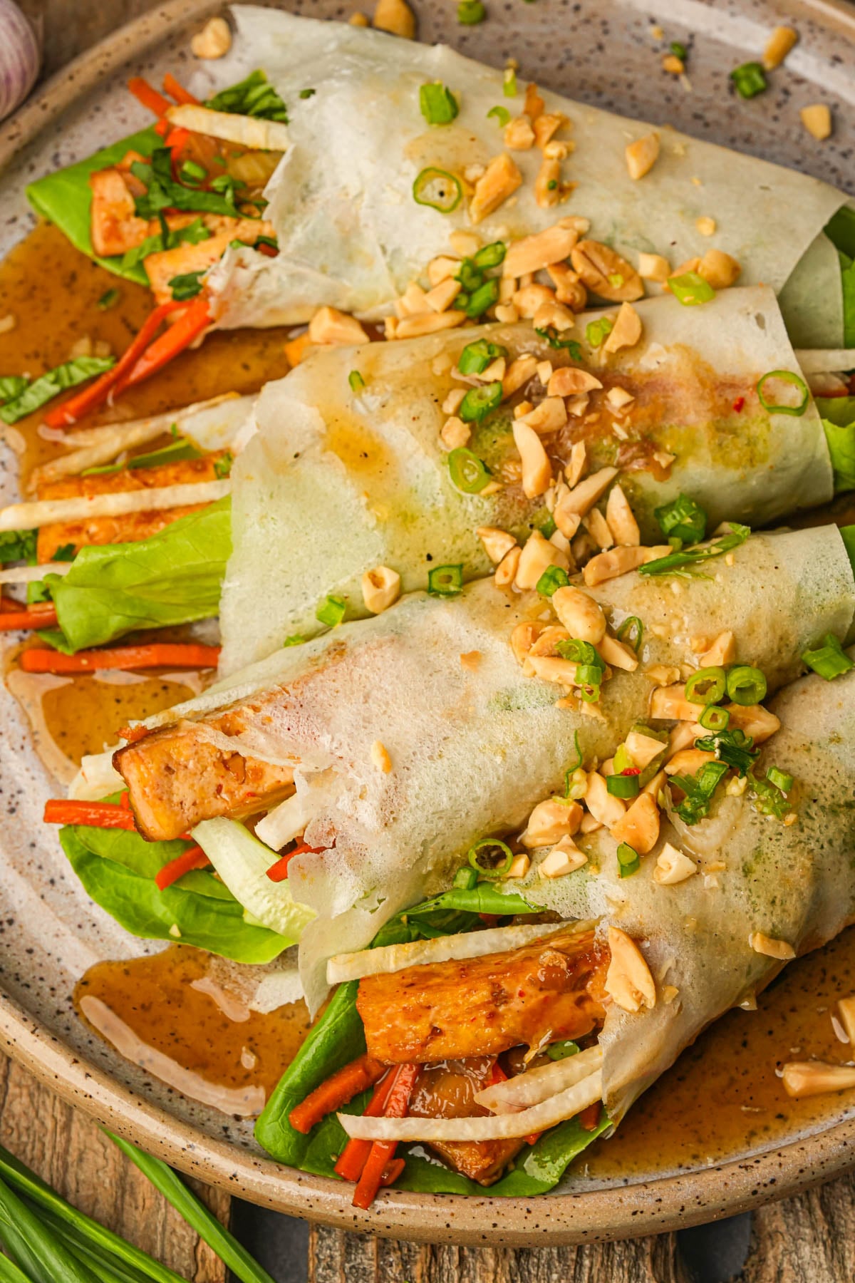 Four fresh lumpiang sariwa rolls filled with tofu, lettuce, carrots, and other vegetables, topped with chopped peanuts, green onions, and a drizzle of sauce, served on a speckled ceramic plate.