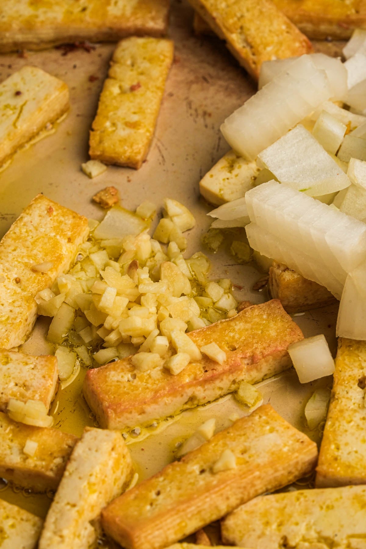 Pan-fried tofu strips with chopped garlic and diced white onions on a skillet, all lightly coated in oil.