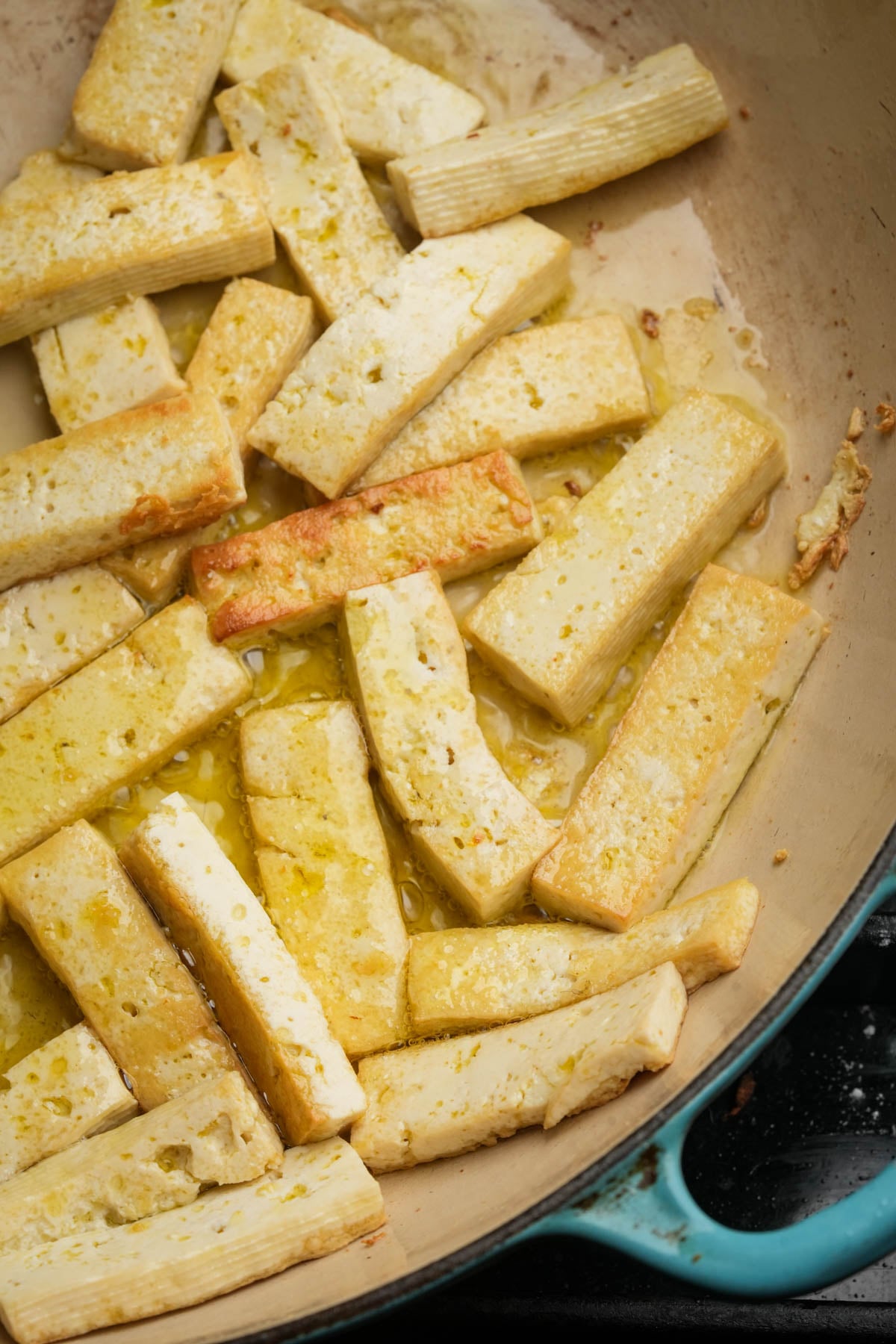 Golden-brown tofu strips are sizzling in a large tan skillet with a blue edge, lightly coated in oil. The tofu appears to be pan-fried and is arranged unevenly across the surface.