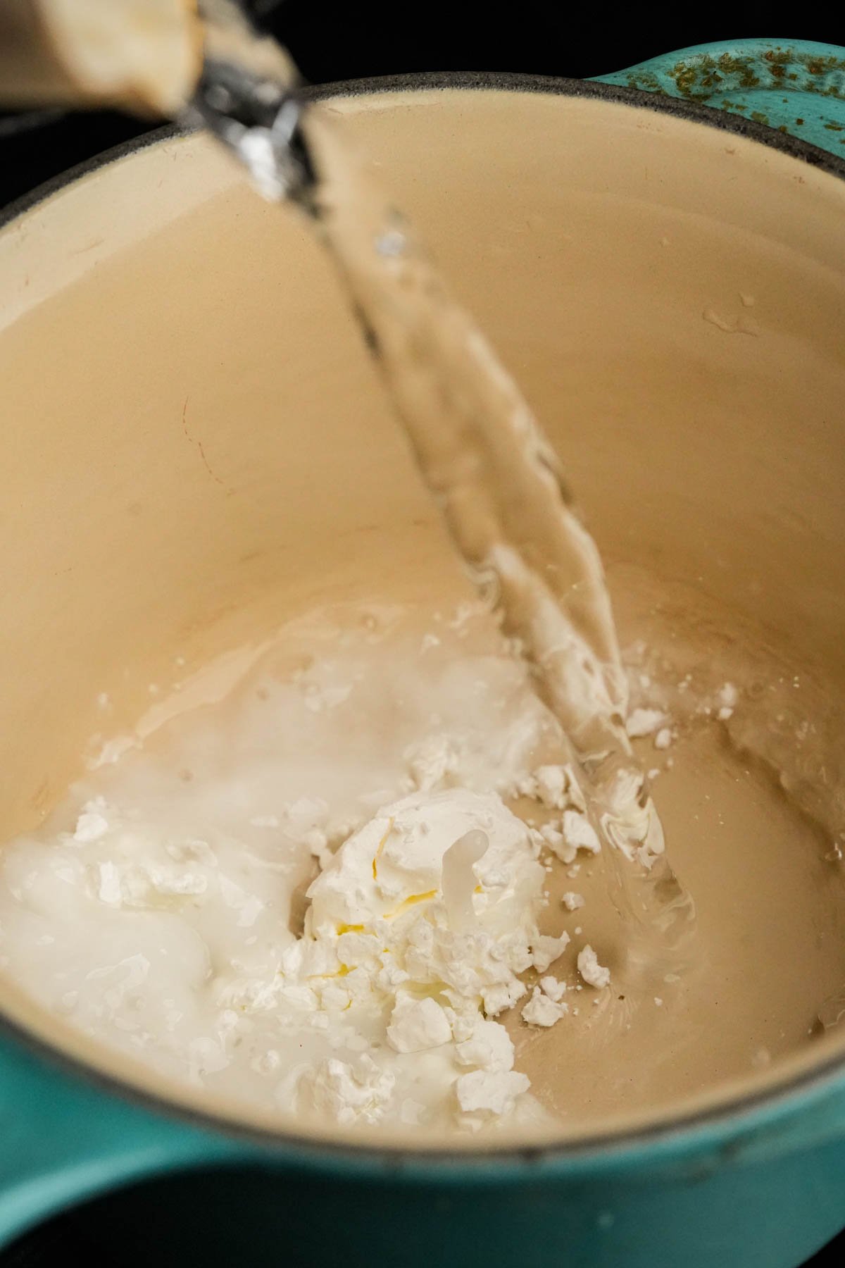 A stream of water is being poured into a light-colored pot containing cornstarch, on a stove. The pot has a turquoise outer edge.