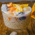 A glass bowl filled with ginataang bilo bilo with tapioca pearls, sweet potato chunks, and purple ube dumplings, jackfruit pieces, set on a wooden surface.