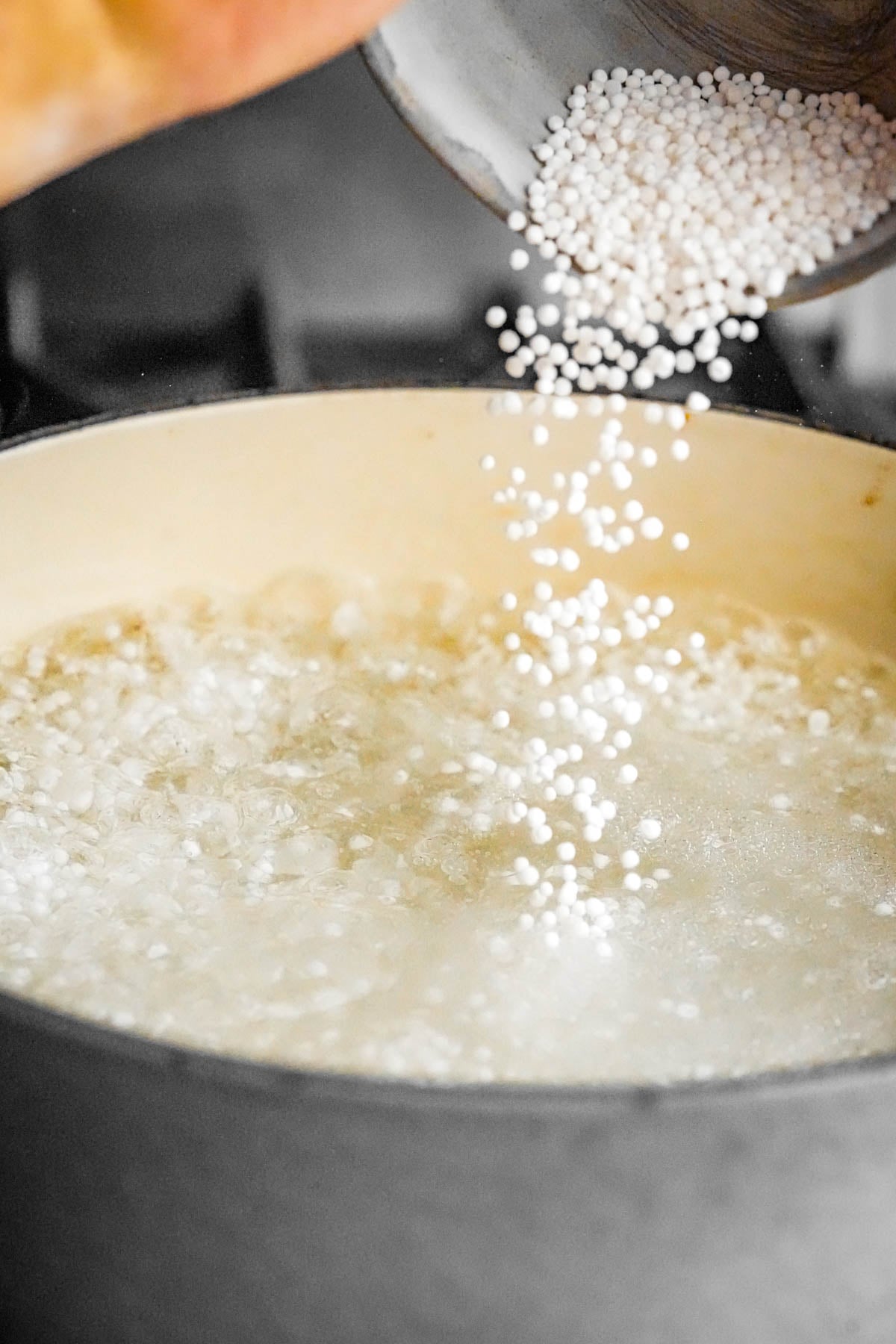 A hand pours small white tapioca pearls from a metal container into a pot of boiling water on a stove.