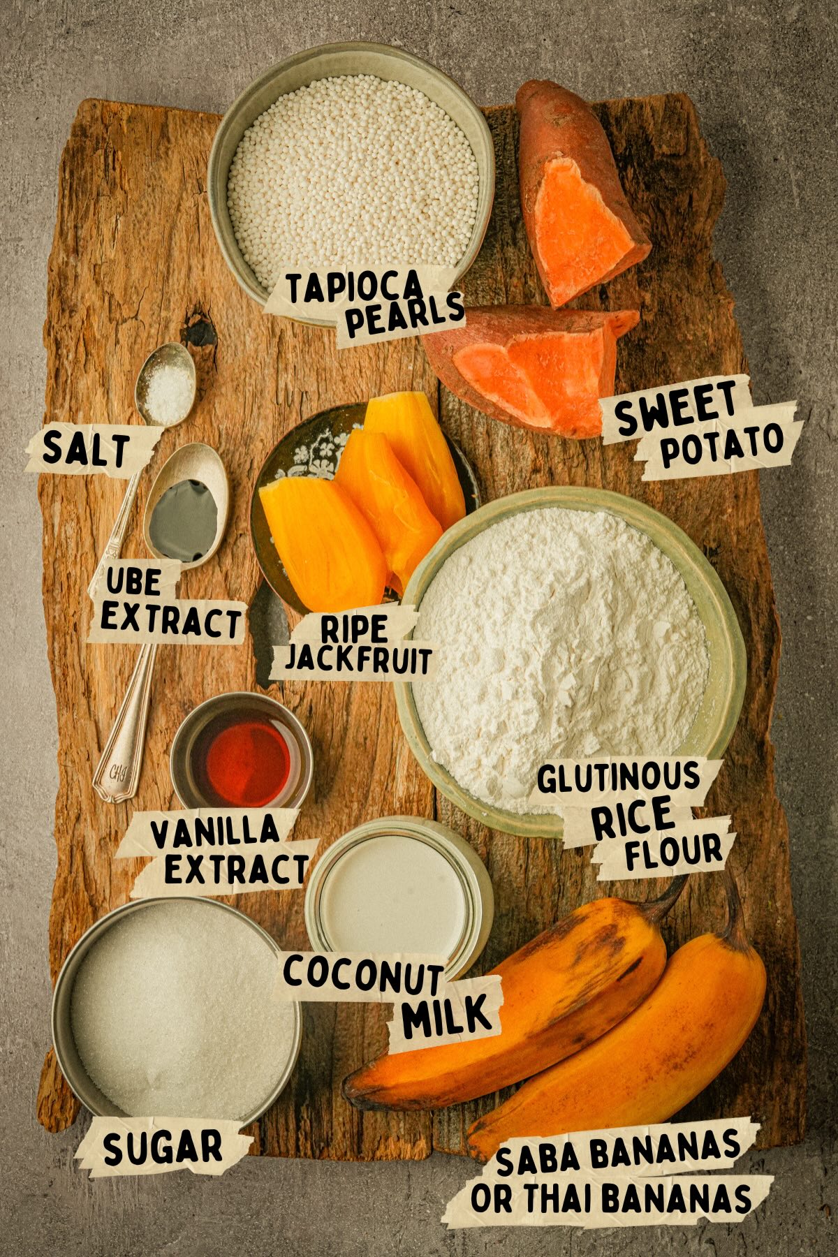 A rustic wooden board displays labeled ingredients for a Filipino dessert, including tapioca pearls, sweet potato, saba bananas, ripe jackfruit, glutinous rice flour, coconut milk, sugar, vanilla extract, ube extract, and salt.