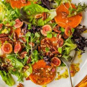 A colorful butternut squash salad with mixed greens, sliced figs, roasted squash rounds, pecans, and a drizzle of dressing, served on a white plate with a spoon.