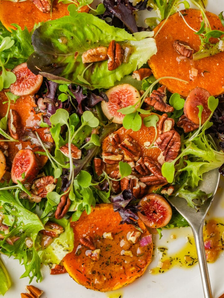 A colorful butternut squash salad with mixed greens, sliced figs, roasted squash rounds, pecans, and a drizzle of dressing, served on a white plate with a spoon.