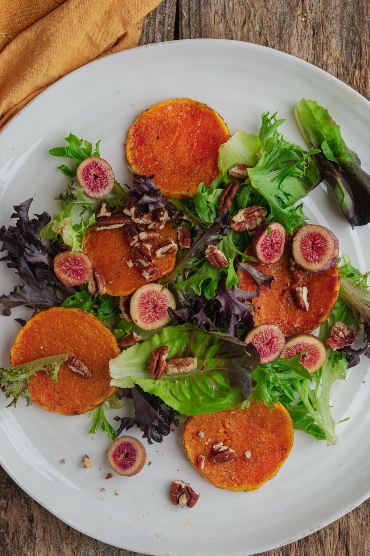 A white plate contains a salad of mixed greens, roasted orange squash rounds, sliced figs, and chopped pecans. The plate sits on a rustic wooden surface with a tan napkin in the corner.