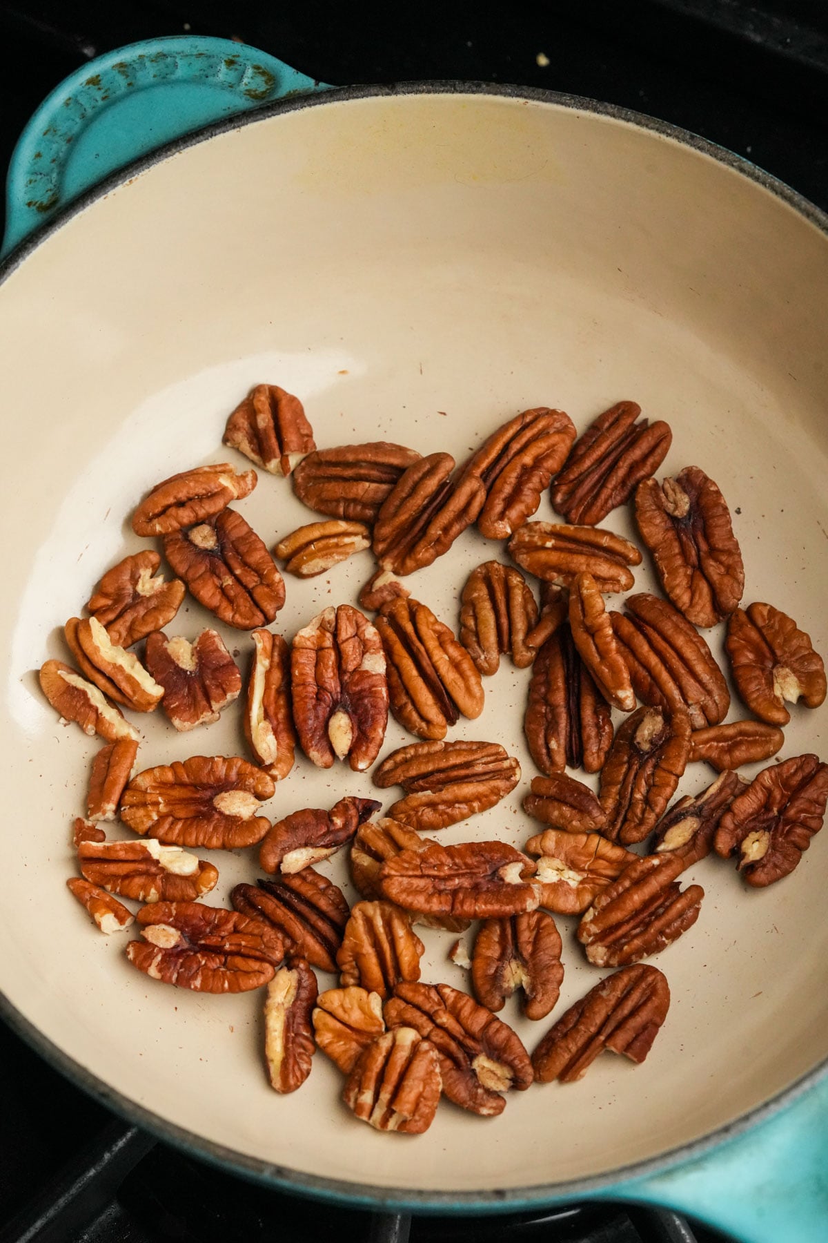 Pecan halves toasting in a cream-colored enameled cast iron pan on a stovetop.