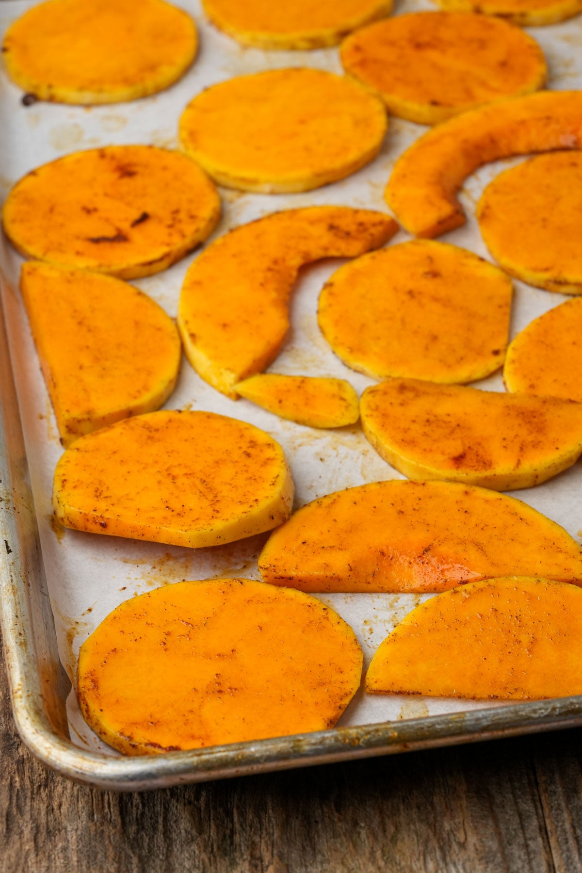 Sliced butternut squash pieces seasoned with spices are arranged on a parchment-lined baking sheet, ready to be roasted.