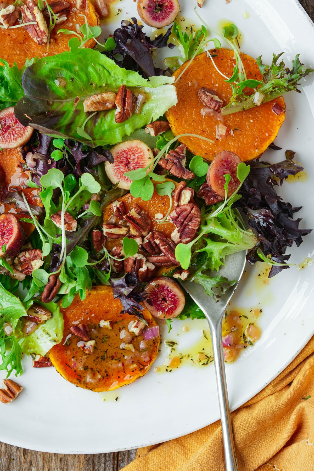A plate of butternut squash salad with mixed greens, sliced figs, roasted squash rounds, pecans, and microgreens, drizzled with vinaigrette; a serving spoon rests on the plate.