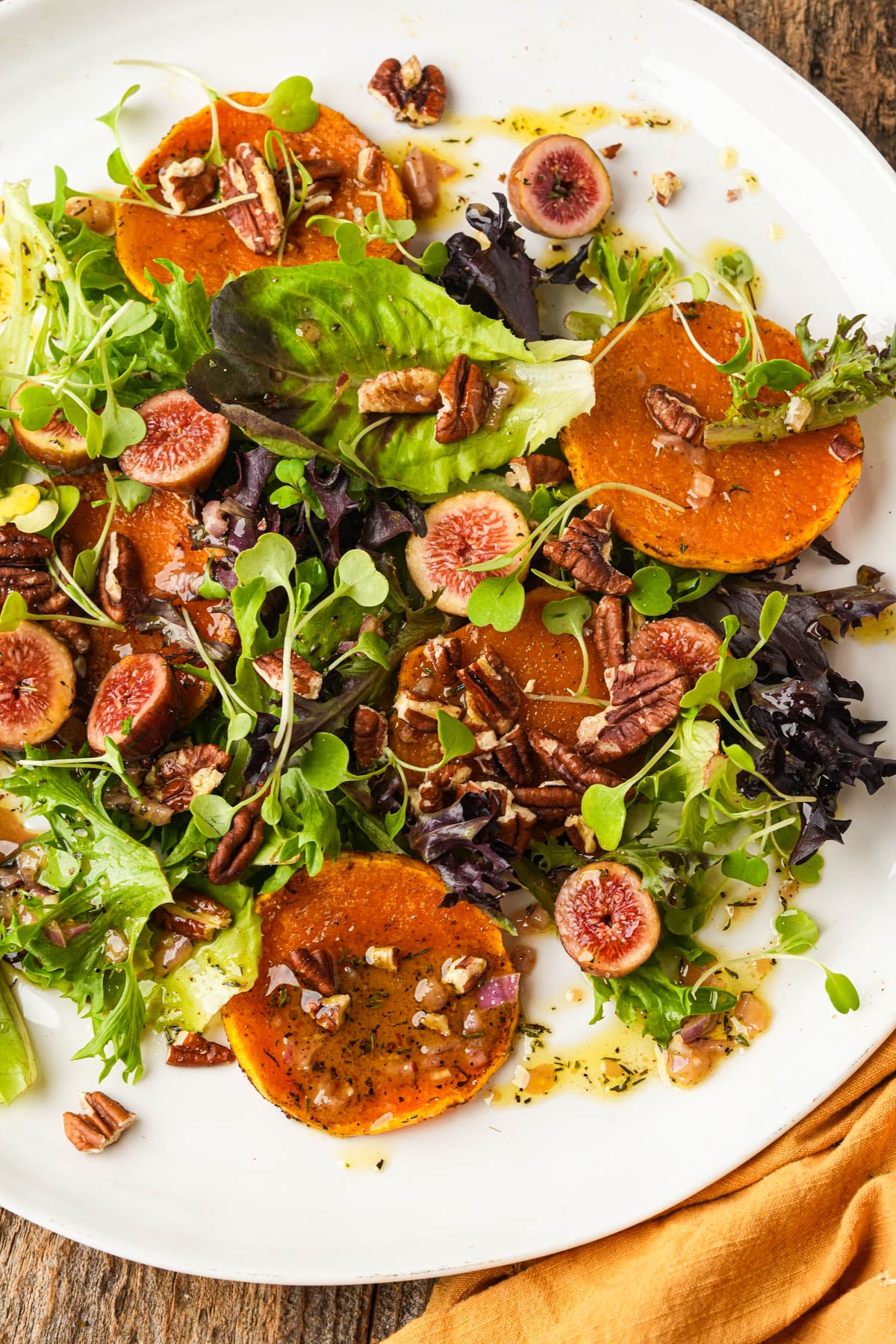A fresh butternut squash salad with mixed greens, roasted squash slices, halved figs, and chopped pecans on a white plate, drizzled with dressing. An orange napkin is partially visible beside the plate.