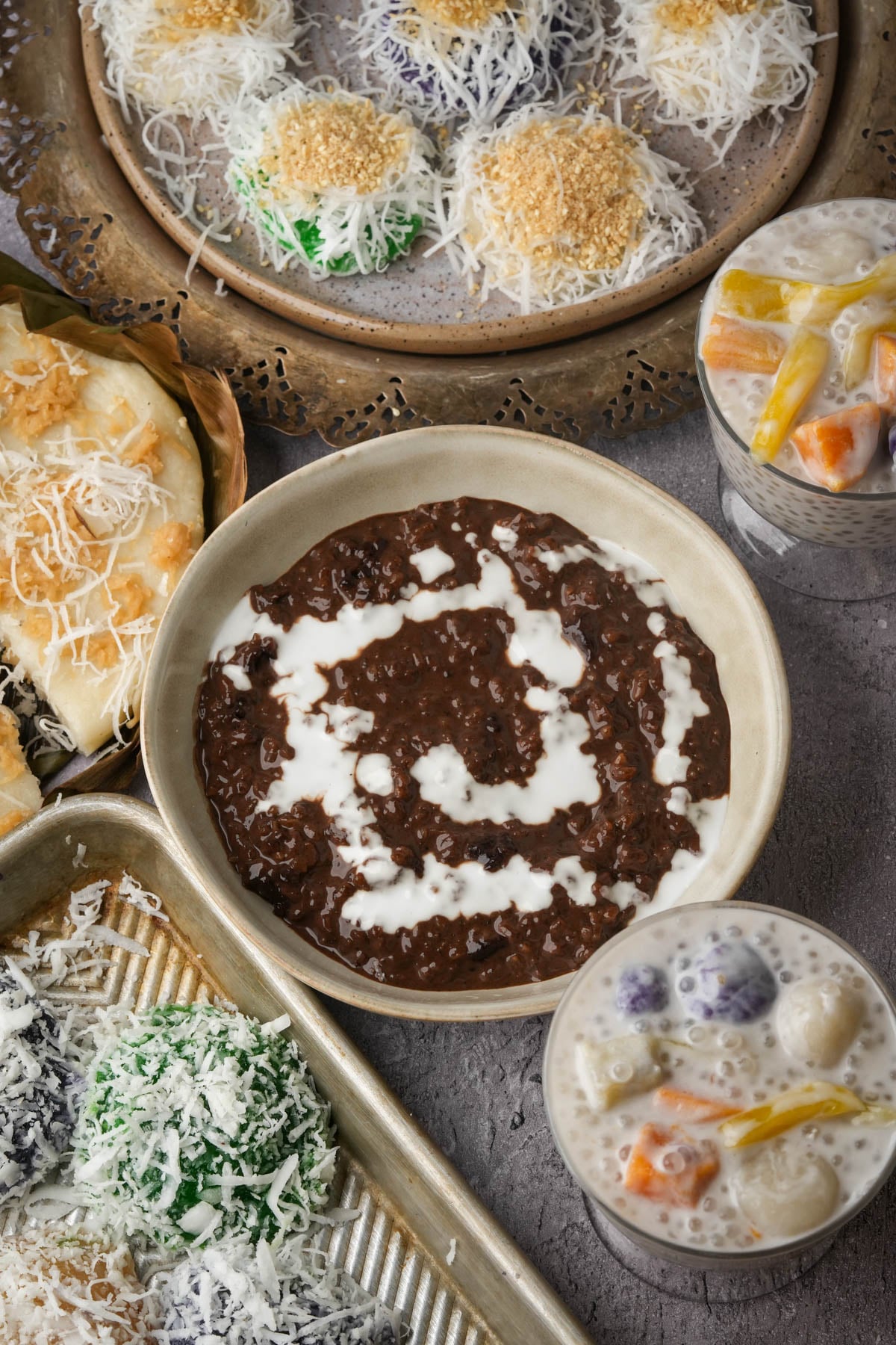 A bowl of vegan champorado with white swirls, assorted Filipino desserts such as pichi pichi and palitaw topped with shredded coconut, bibingka, and two glasses of creamy bilo bilo are arranged on a table.