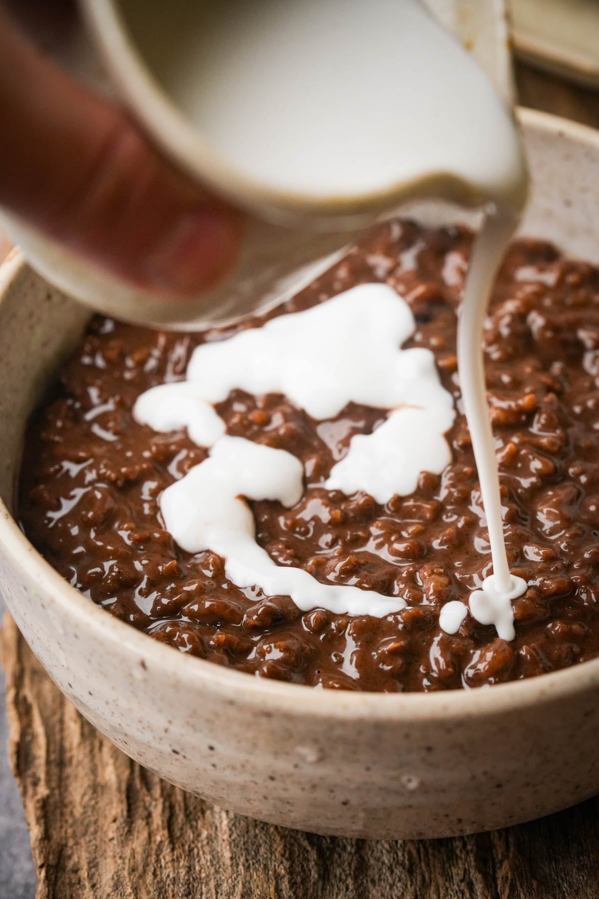 A close-up of vegan champorado in a ceramic bowl, with a hand pouring white coconut milk over it from a small pitcher. The bowl sits on a rustic wooden surface.