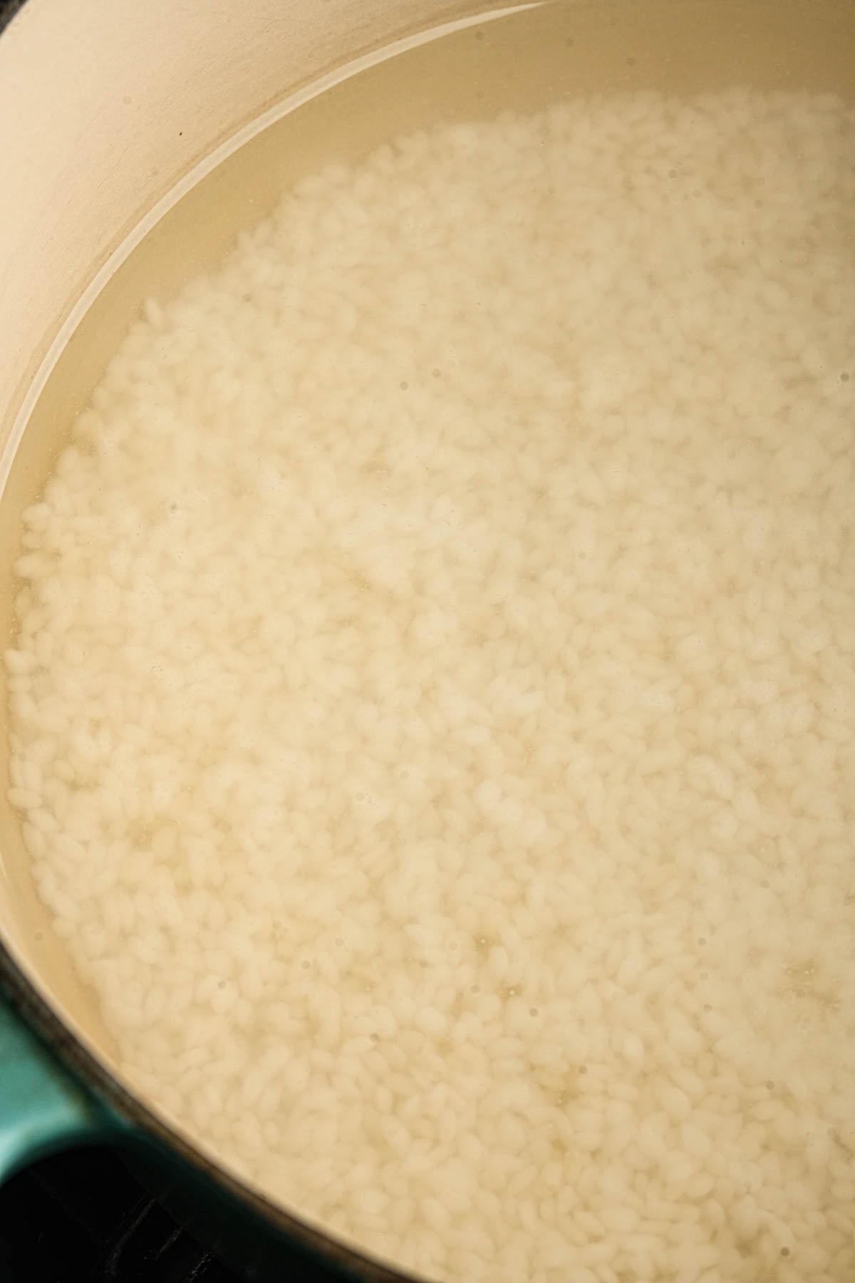 A close-up of uncooked sweet rice cooking in water inside a large, light-colored pot.