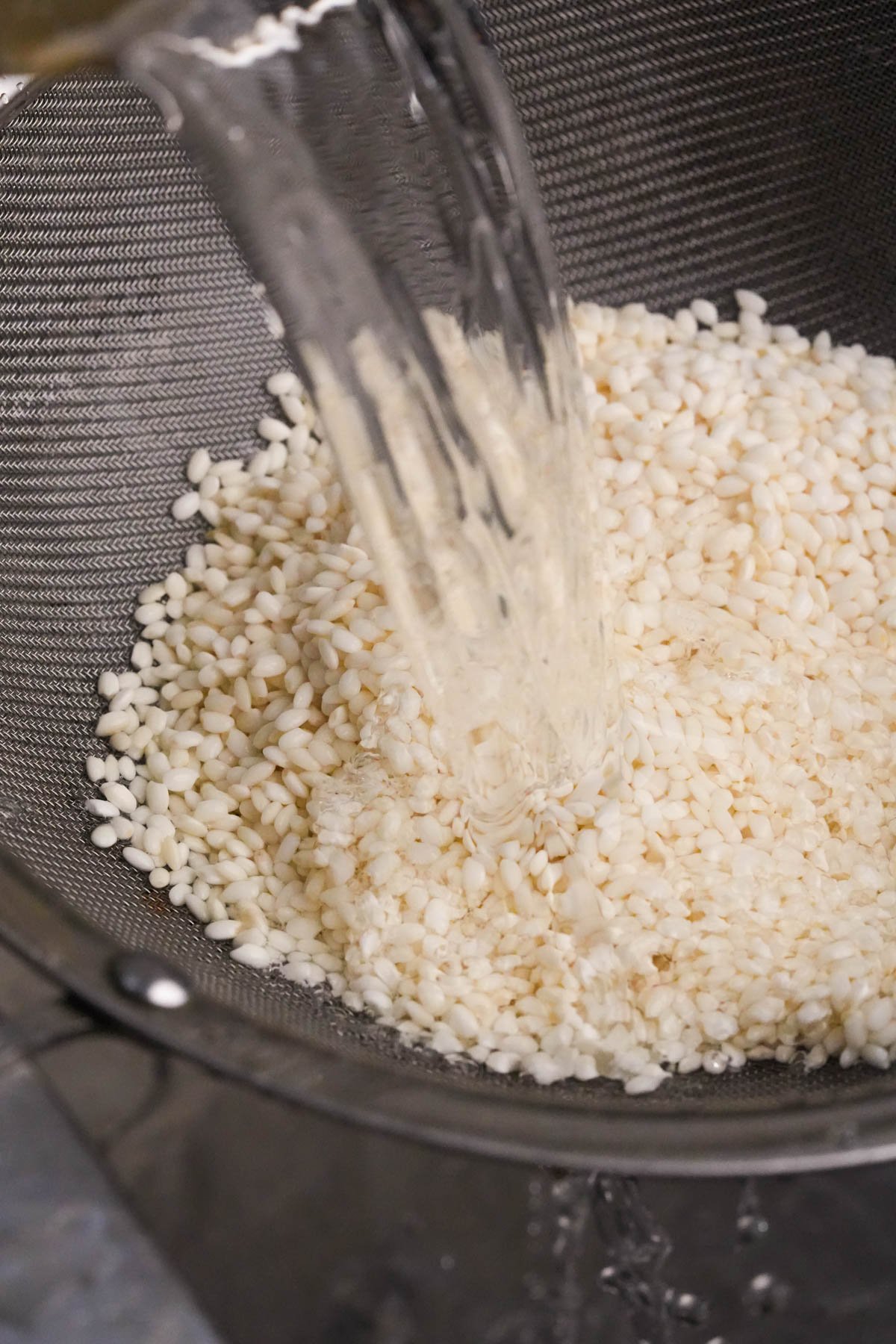 Water is being poured over sweet rice grains in a metal mesh strainer, rinsing them before cooking.