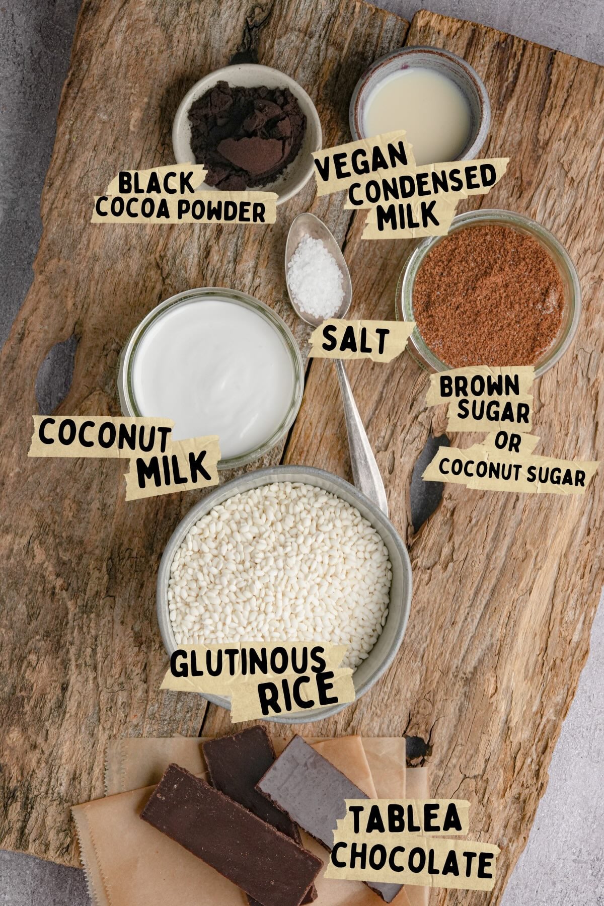 A wooden surface displays bowls of ingredients for a champorado recipe including black cocoa powder, vegan condensed milk, coconut milk, salt, brown or coconut sugar, glutinous rice, and pieces of tablea chocolate, each labeled with handwritten tags.