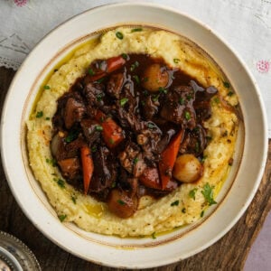 A bowl of creamy mashed potatoes topped with mushroom bourguignon containing chunks of mushroom, carrots, pearl onions, and fresh herbs, garnished with chopped chives.
