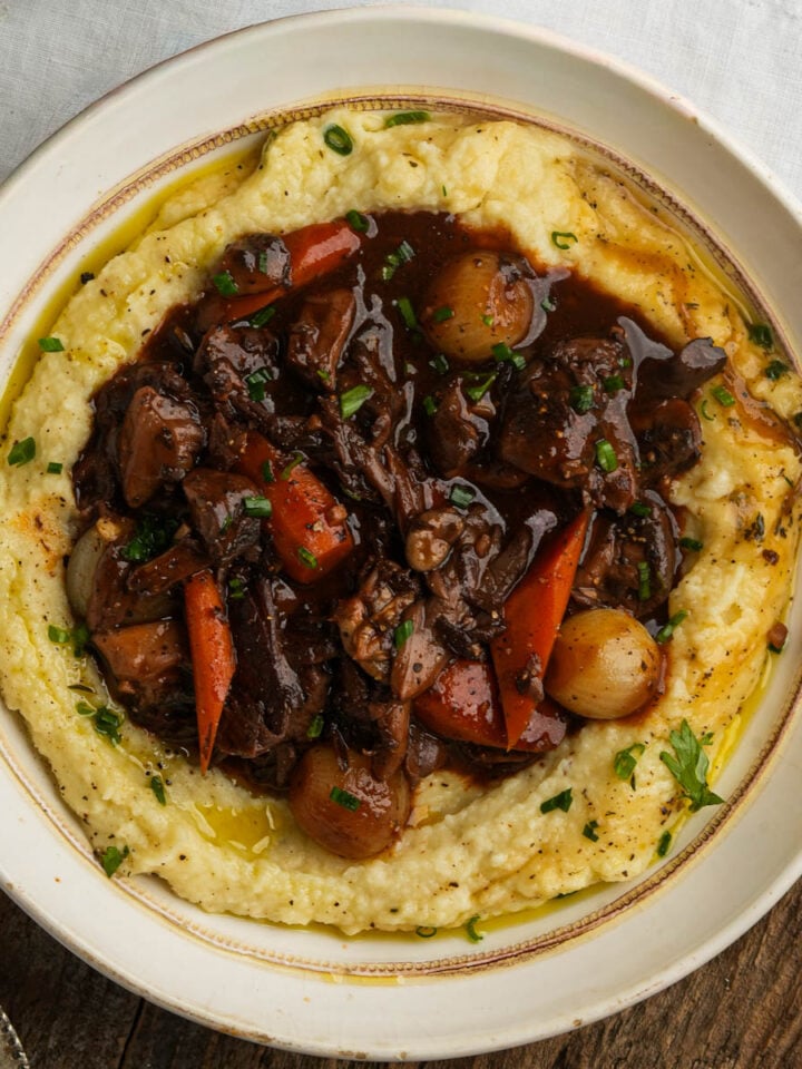 A bowl of creamy mashed potatoes topped with mushroom bourguignon containing chunks of mushroom, carrots, pearl onions, and fresh herbs, garnished with chopped chives.