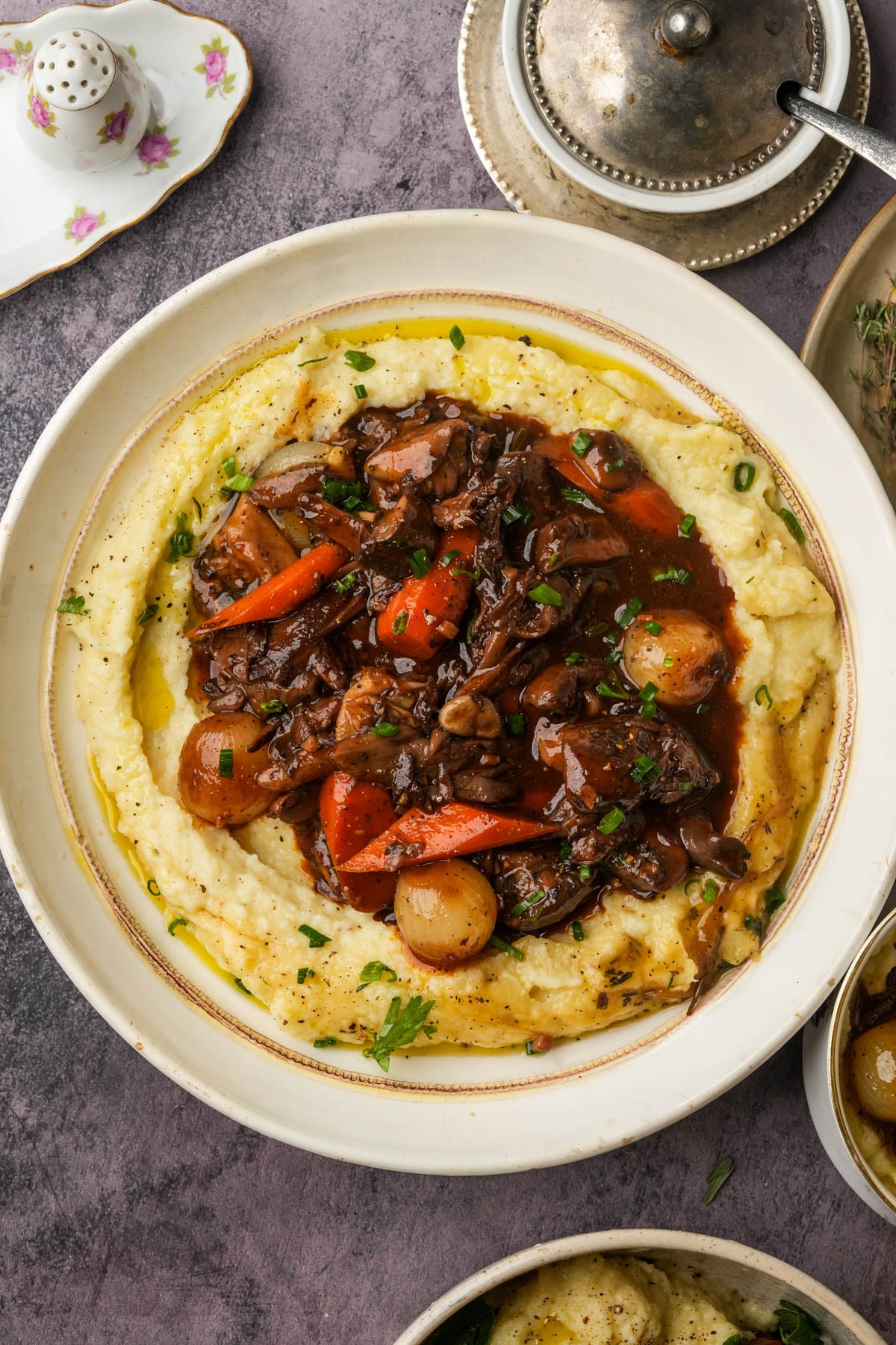 A bowl of creamy mashed potatoes topped with a mushroom bourguignon containing carrots, pearl onions, and fresh herbs, served on a rustic tabletop with dishes and a pepper shaker nearby.