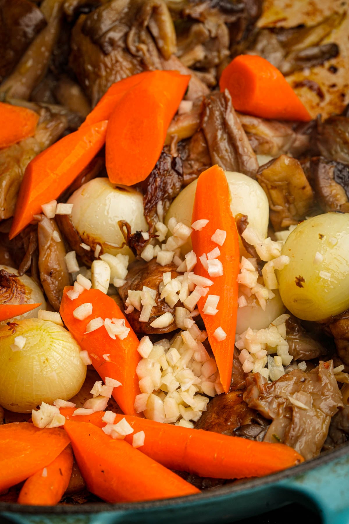 Close-up of a dish featuring braised carrots, pearl onions, chopped garlic, and tender pieces of mushroom, all cooked together, with vibrant orange and golden tones.