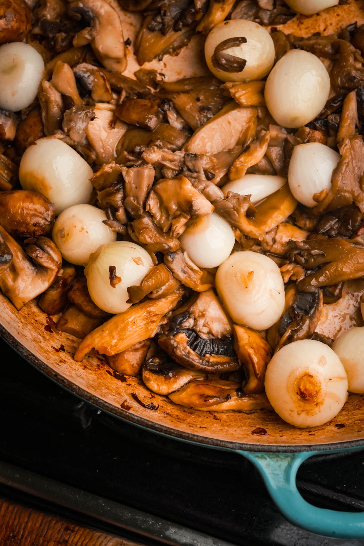 A pan filled with sautéed mushrooms and whole pearl onions, cooked to a golden brown. The dish is shown in a blue-edged skillet on a stovetop.