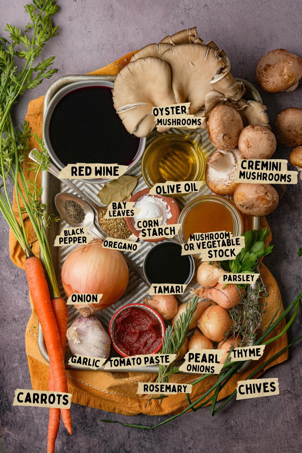 An overhead view of labeled ingredients for a mushroom bourguignon recipe including red wine, oyster and cremini mushrooms, olive oil, bay leaves, cornstarch, tamari, carrots, onions, garlic, tomato paste, herbs, and seasonings.