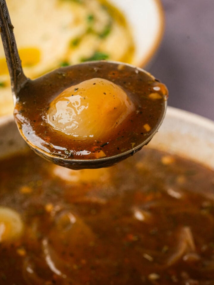 A ladle holding a serving of onion gravy. A plate of mashed potatoes is blurred in the background.