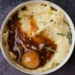 A bowl of creamy mashed potatoes topped with brown onion gravy, a whole cooked onion, black pepper, and fresh rosemary on a dark surface.