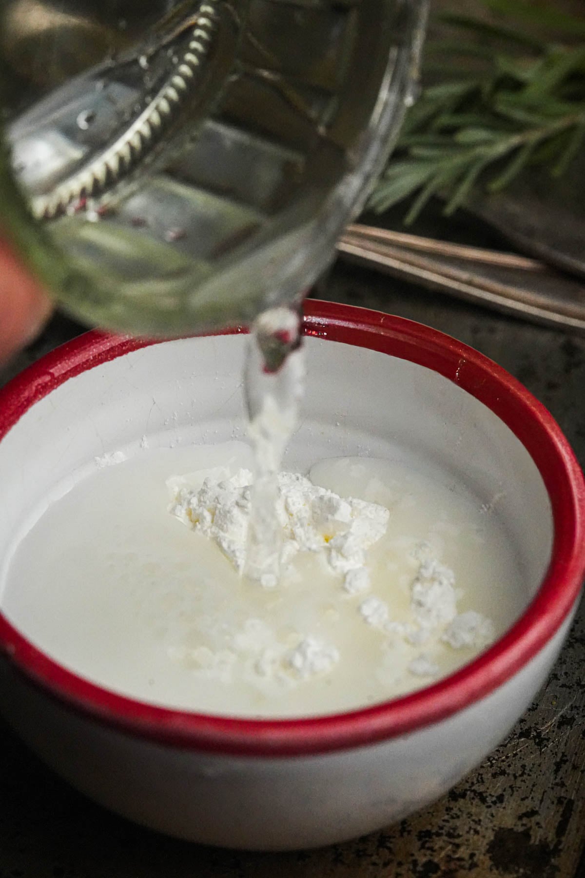 A close-up of a red-rimmed bowl containing white liquid and cornstarch, with water being poured in from a glass above. Green herbs are blurred in the background.