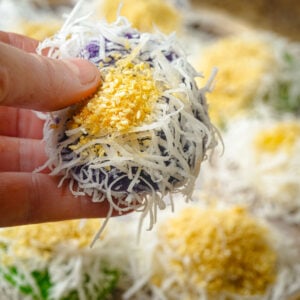 A hand holding a colorful Filipino rice cake, palitaw, coated with shredded coconut and sprinkled with sesame seeds and sugar, with more rice cakes blurred in the background.