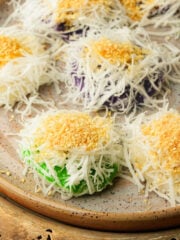 A plate of palitaw, Filipino steamed rice cakes in pandan, ube, and plain, topped with shredded coconut and toasted sesame sugar on a speckled ceramic dish.