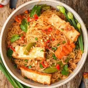A bowl of stir-fried pancit bihon noodles with slices of tofu, bell peppers, carrots, chopped green onions, cilantro, and red chili peppers, garnished with fresh herbs.