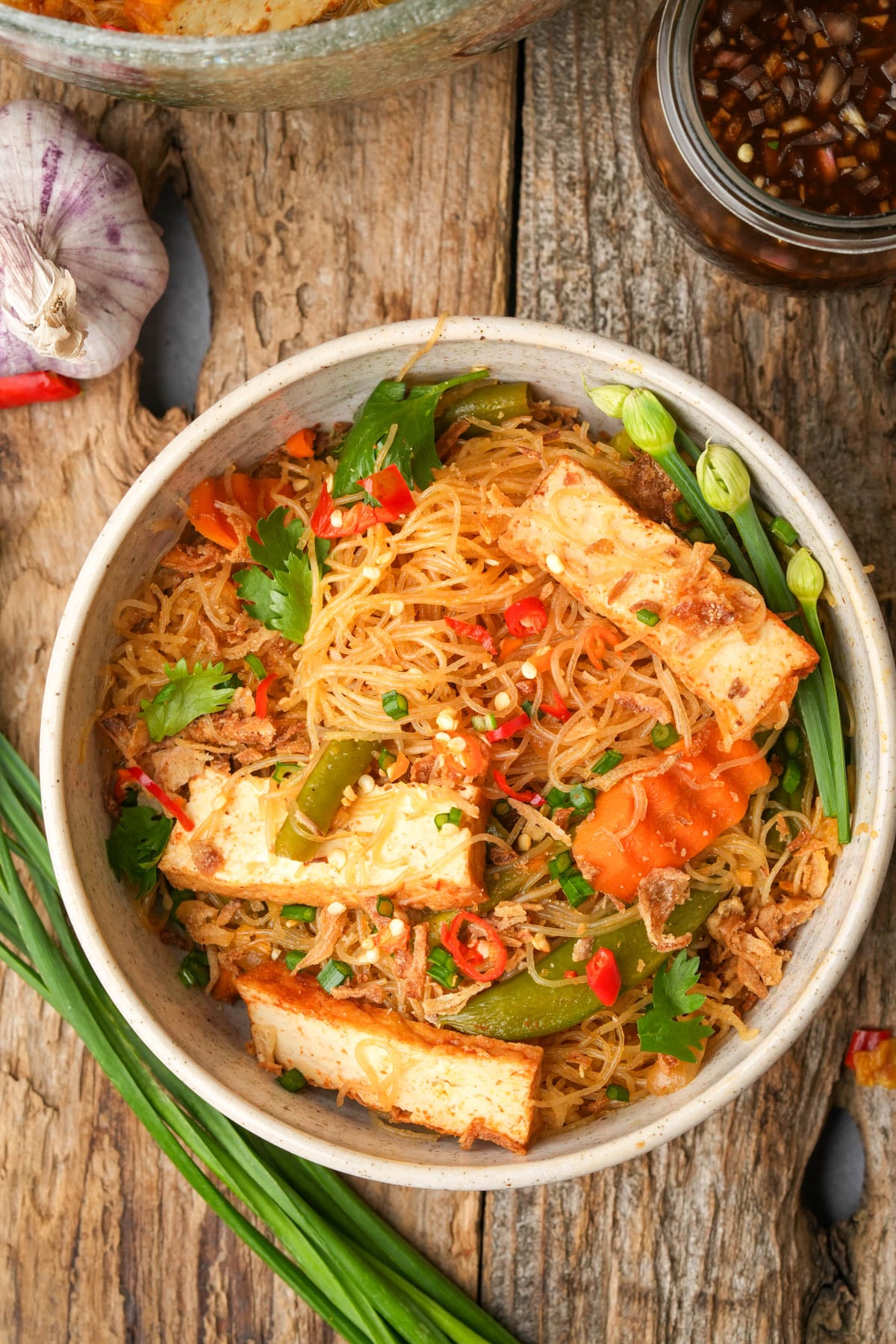 A bowl of stir-fried pancit bihon noodles with tofu strips, sliced vegetables, chopped green onions, fresh cilantro, and red chili pepper, garnished with crushed peanuts, placed on a rustic wooden table.