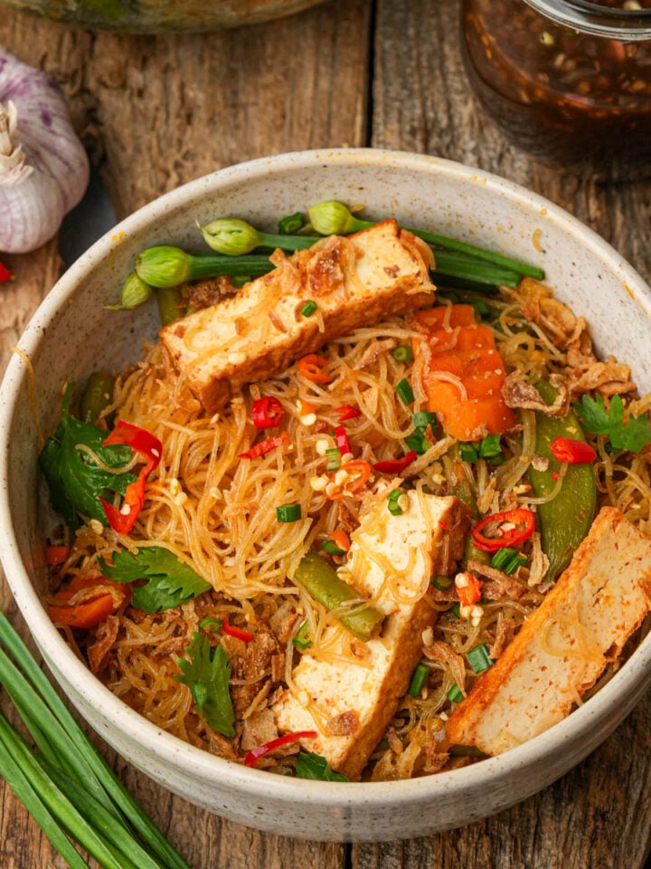 A bowl of stir-fried pancit bihon noodles with vegetables, sliced tofu, red chili, green onions, and fresh herbs, placed on a rustic wooden table. Garlic, red chili, and a jar of sauce are nearby.