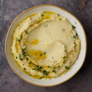A bowl of creamy parsnip puree topped with olive oil, chopped chives, and cracked black pepper, set on a dark textured surface.