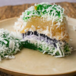 A slice of colorful Filipino pichi pichi cake topped with shredded coconut, showing layers of green, purple, and yellow, on a beige plate with a rustic wooden background.