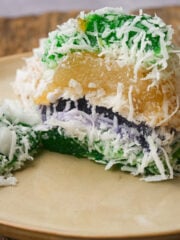 A slice of colorful Filipino pichi pichi cake topped with shredded coconut, showing layers of green, purple, and yellow, on a beige plate with a rustic wooden background.
