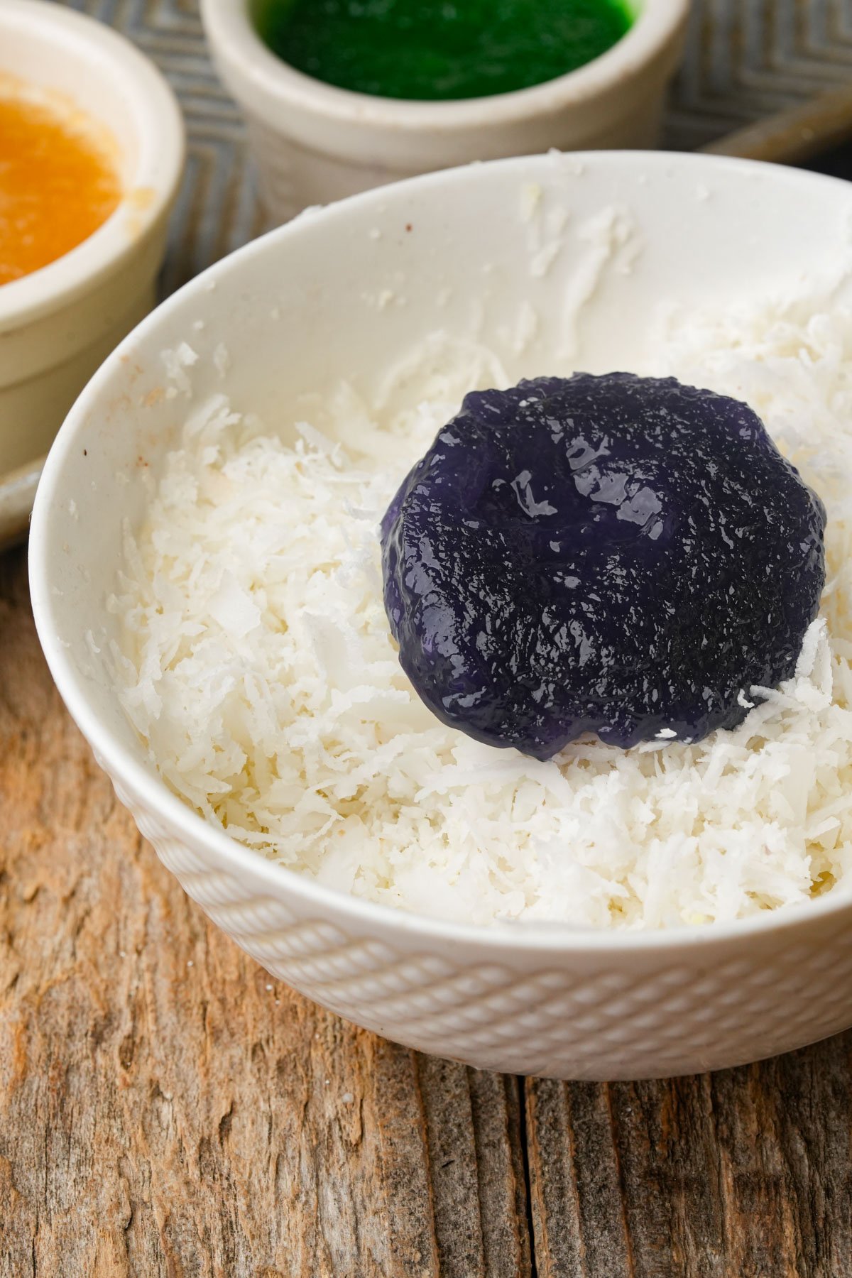 A white bowl filled with shredded coconut and topped with a round, purple jelly-like pichi pichi. In the background, small bowls with green and yellow pichi pichi are visible on a wooden surface.