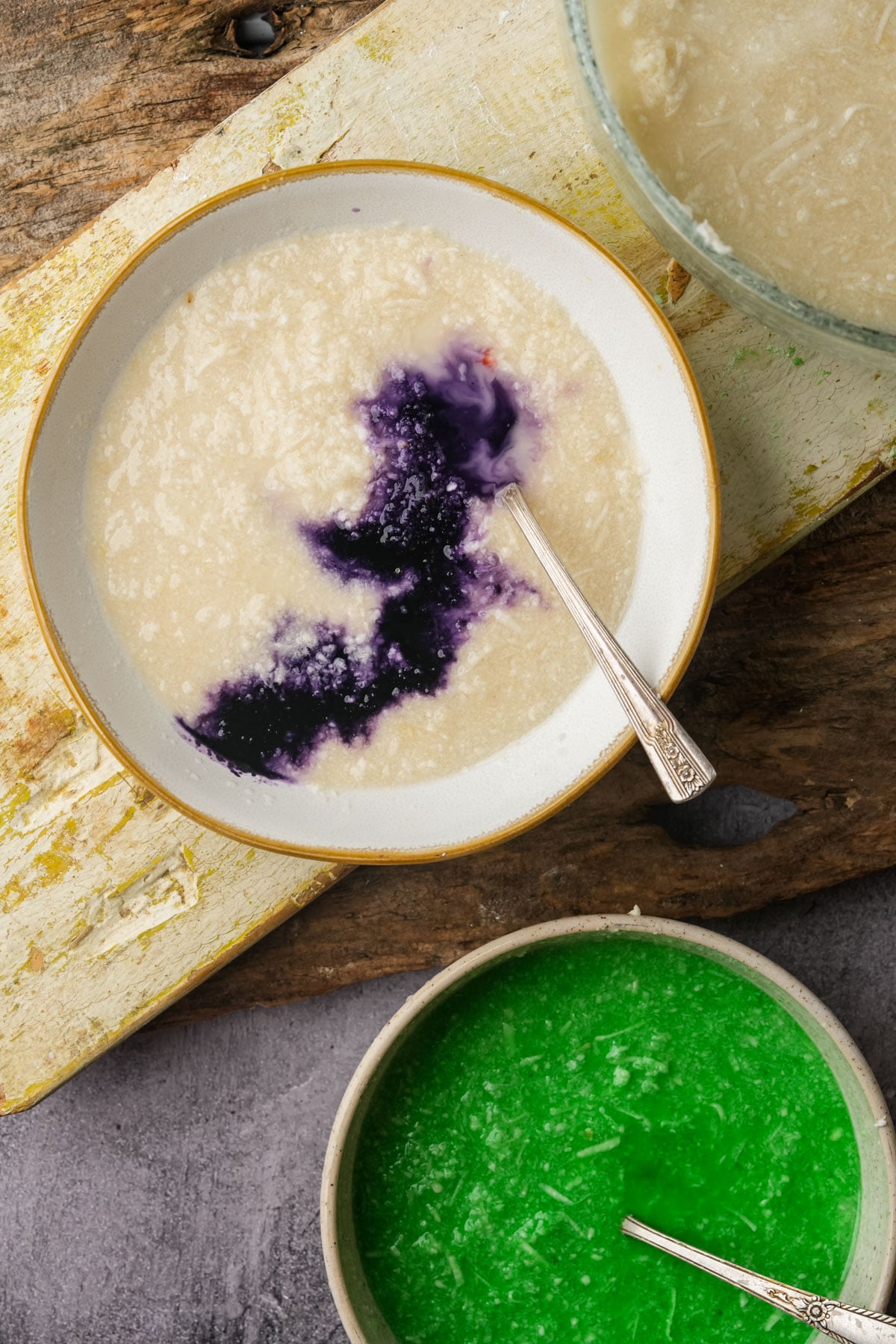 A bowl of creamy white pichi pichi batter with a splash of dark purple ube extract, a silver spoon resting inside. Nearby are bowls of pandan mixed batter and plain batter on a rustic wooden surface.