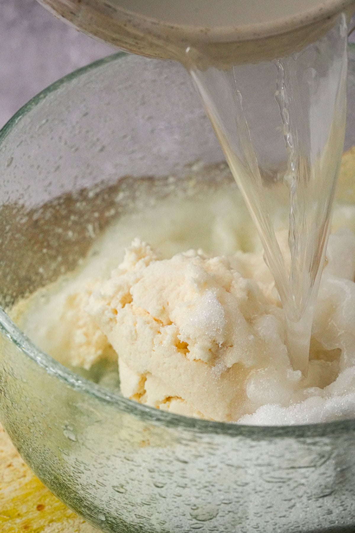 A close-up of a glass bowl containing a mound of white rice flour or sugar, with water being poured over it from a container above.