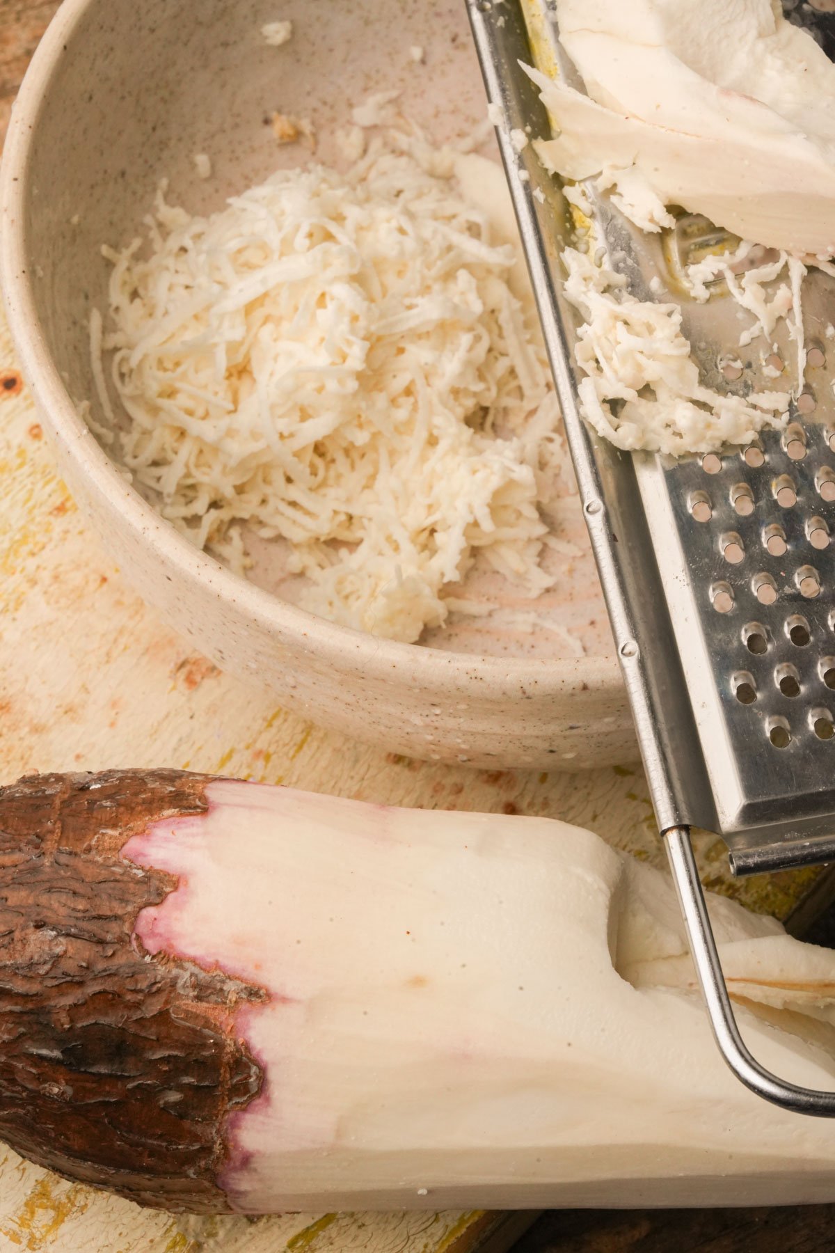 A grated cassava root sits in a beige bowl next to a metal grater, with a whole, partially peeled root on a wooden cutting board beside them.