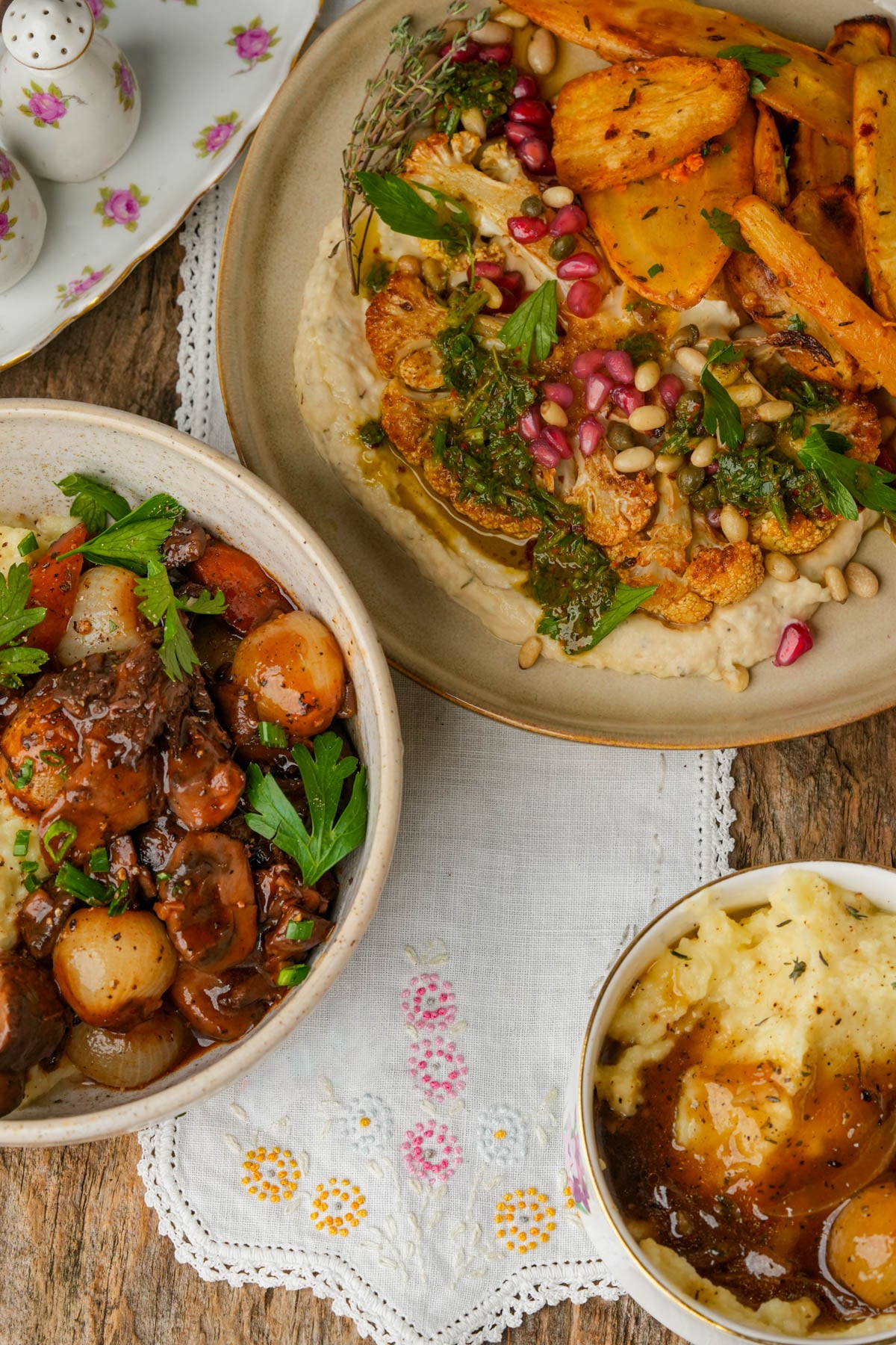 A table with three dishes: mashed potatoes with onion gravy, a bowl of mushroom bourguignon with pearl onions and parsley, and roasted cauliflower steak on bean puree topped with pine nuts, pomegranate seeds, roasted parsnips and herbs on a beige plate.