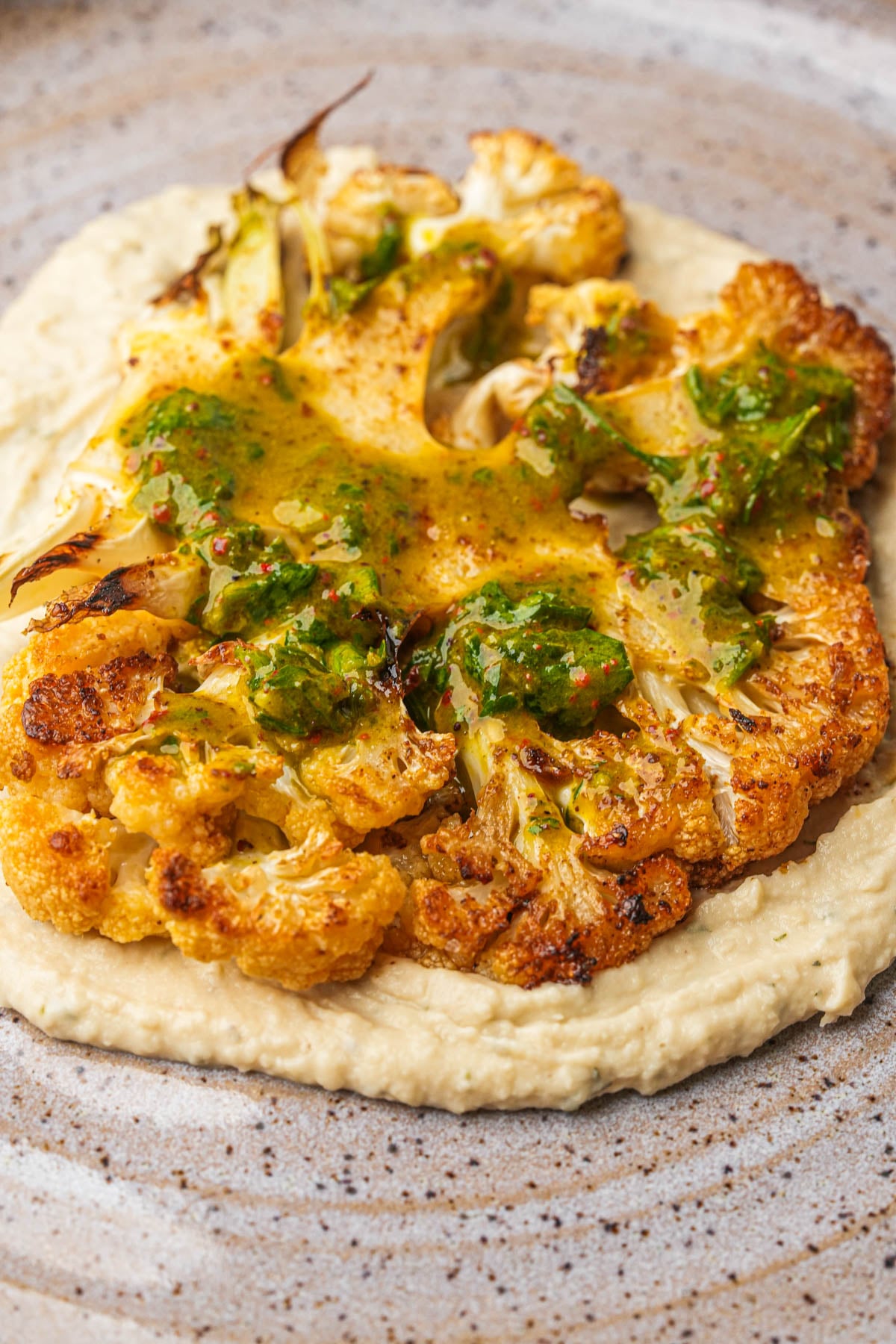 A roasted cauliflower steak sits on a bed of creamy bean puree, topped with parsley sauce, served on a speckled ceramic plate.