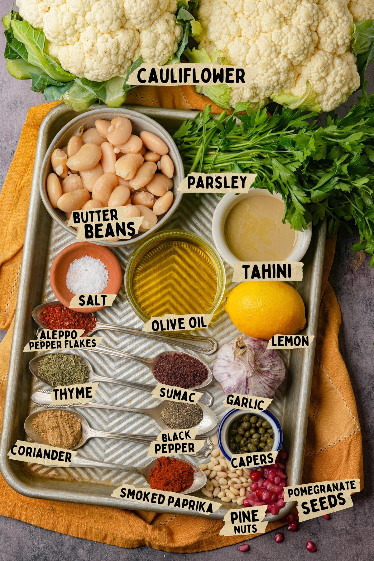 A tray displays ingredients for a roasted cauliflower steak recipe: cauliflower, butter beans, parsley, tahini, olive oil, lemon, garlic, capers, pomegranate seeds, pine nuts, and spices such as salt, thyme, coriander, sumac, paprika, black pepper, and Aleppo pepper flakes.