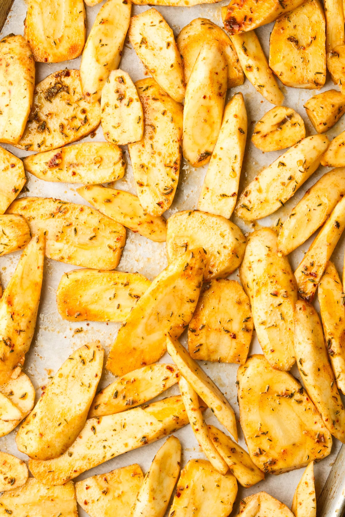 Thin slices of seasoned parsnips spread out on a baking sheet, ready for roasting. The slices are coated with herbs and spices, giving them a golden, slightly speckled appearance.