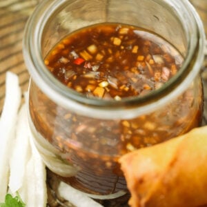 A glass jar filled with toyomansi dipping sauce sits on a wooden surface, next to a spring roll and thin slices of white vegetable.