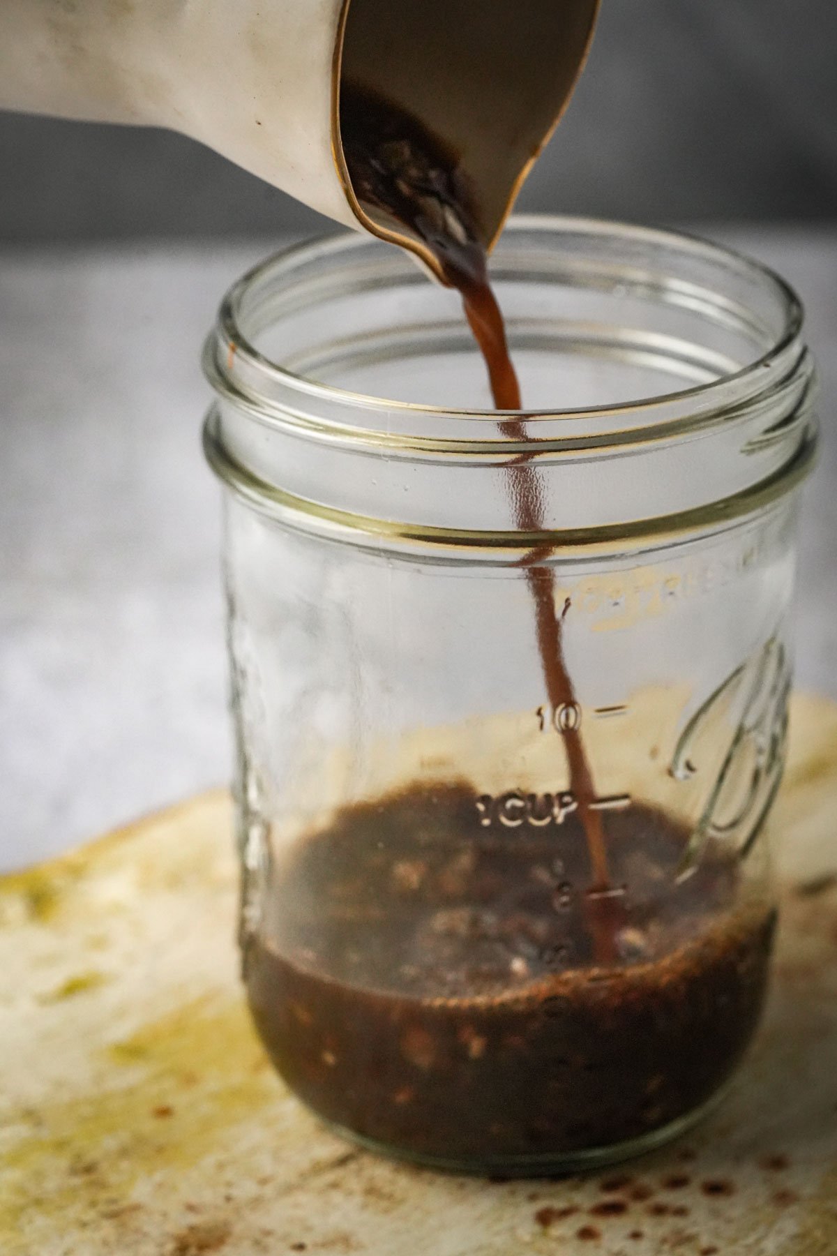 A stream of toyomansi is being poured from a small pitcher into a clear glass mason jar, which sits on a light-colored surface. Some of the liquid has already collected in the jar.