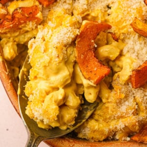 Close-up of creamy vegan butternut squash macaroni and cheese topped with golden-brown roasted squash and a crispy breadcrumb crust, with a serving spoon lifting a portion from the dish.