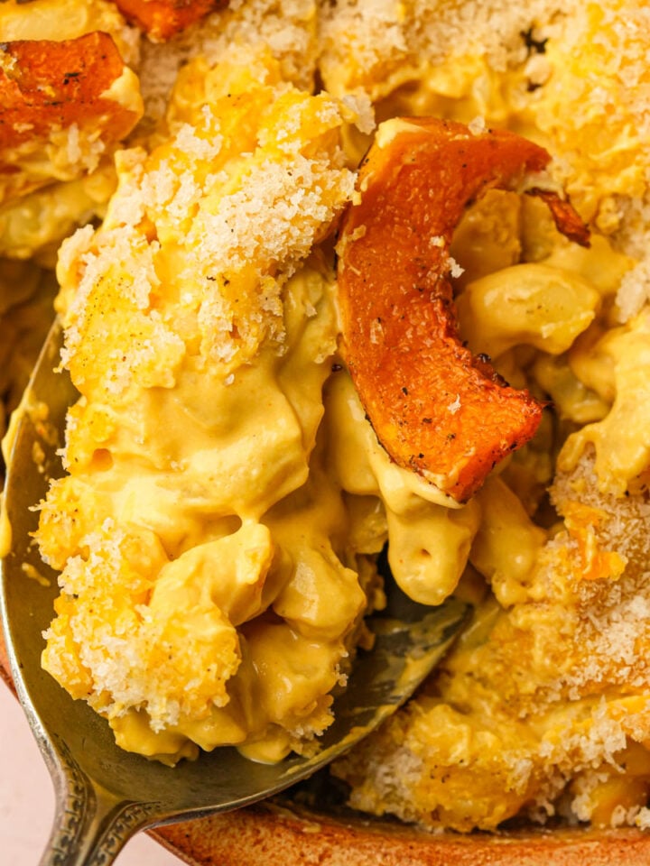 Close-up of creamy vegan butternut squash macaroni and cheese topped with golden-brown roasted squash and a crispy breadcrumb crust, with a serving spoon lifting a portion from the dish.